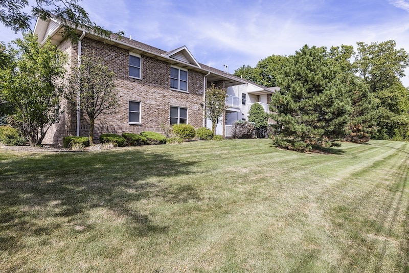 Brick building with white siding on a grassy lawn, surrounded by trees and shrubs under a clear sky.