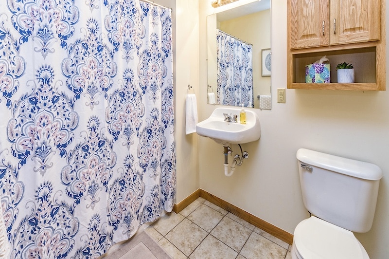 Bathroom with blue floral shower curtain, wall-mounted sink, a mirror, wooden cabinet, toilet, and beige tiled floor.