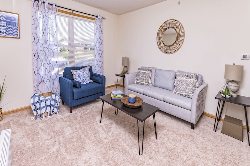 A living room with a blue armchair, gray sofa, and black coffee table. Decor includes blue and white cushions, a wall mirror, two lamps, sheer curtains, and framed artwork.