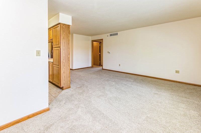 Empty room with beige carpet and white walls, featuring a wood-trimmed corner and an open doorway leading to another room.