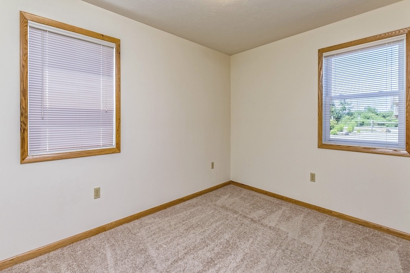 Empty room with beige carpet and white walls, featuring two windows with blinds and wooden trim.