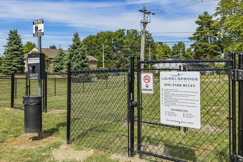 Entrance to a dog park with a sign displaying park rules and amenities. A waste station and "No Smoking" sign are visible on the fence.