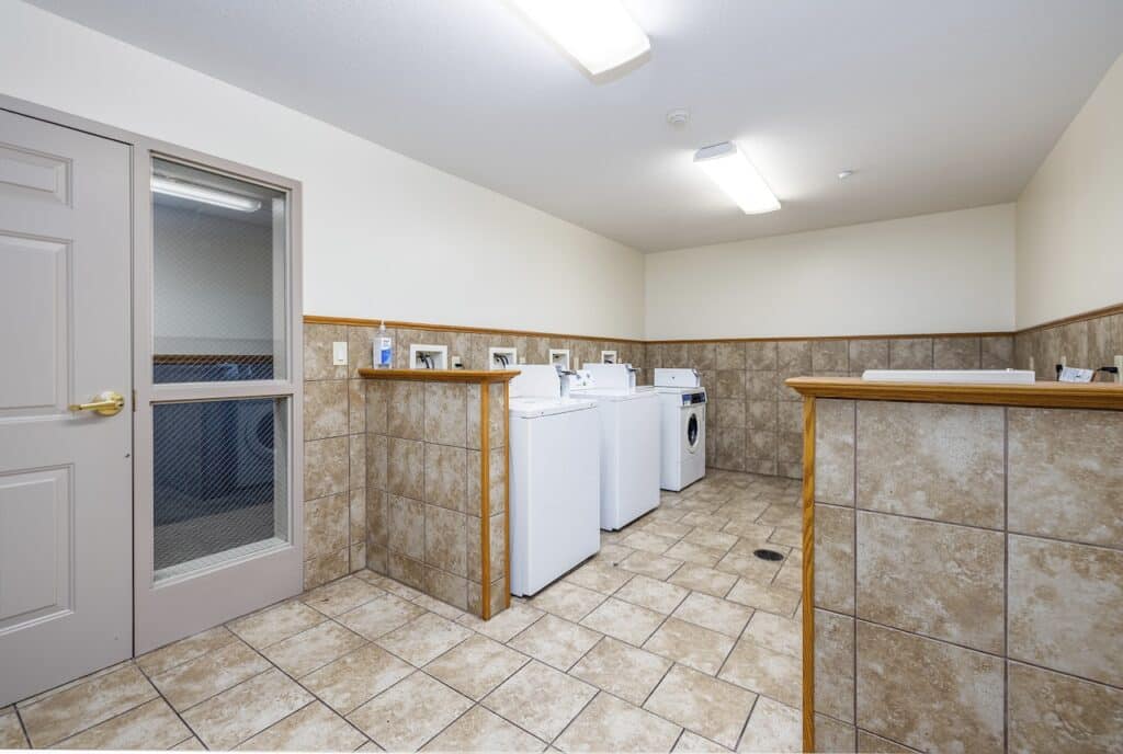 Laundry room with tiled walls and floor, featuring several washing machines and dryers, under fluorescent lighting.