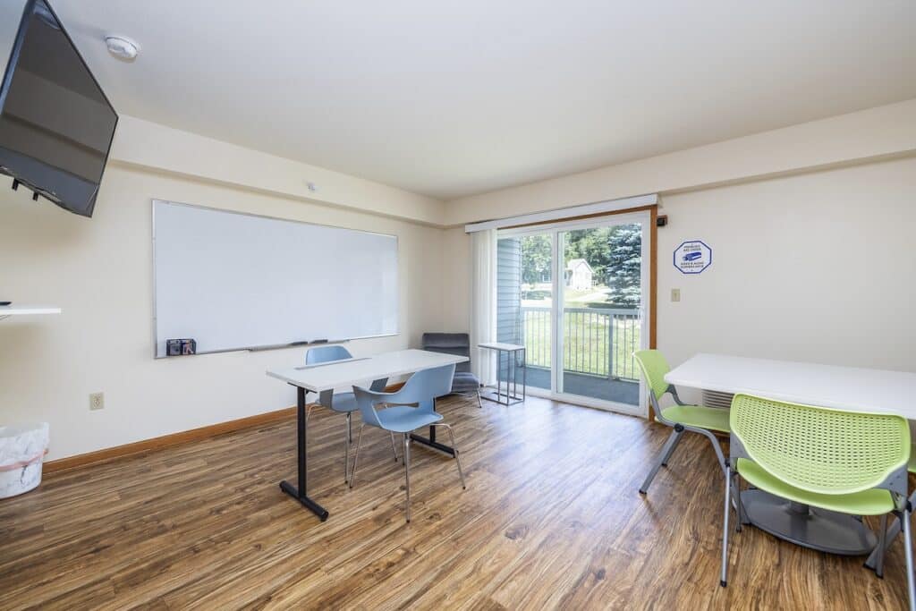 Small classroom with wooden flooring, two tables, assorted chairs, a whiteboard, wall-mounted TV, and a glass door leading outside.