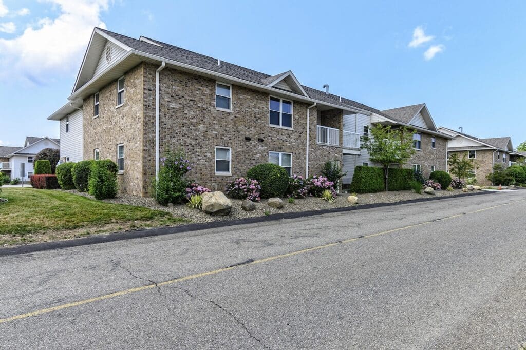 A two-story apartment building with brick and siding exterior, surrounded by shrubs and a paved road in the foreground.