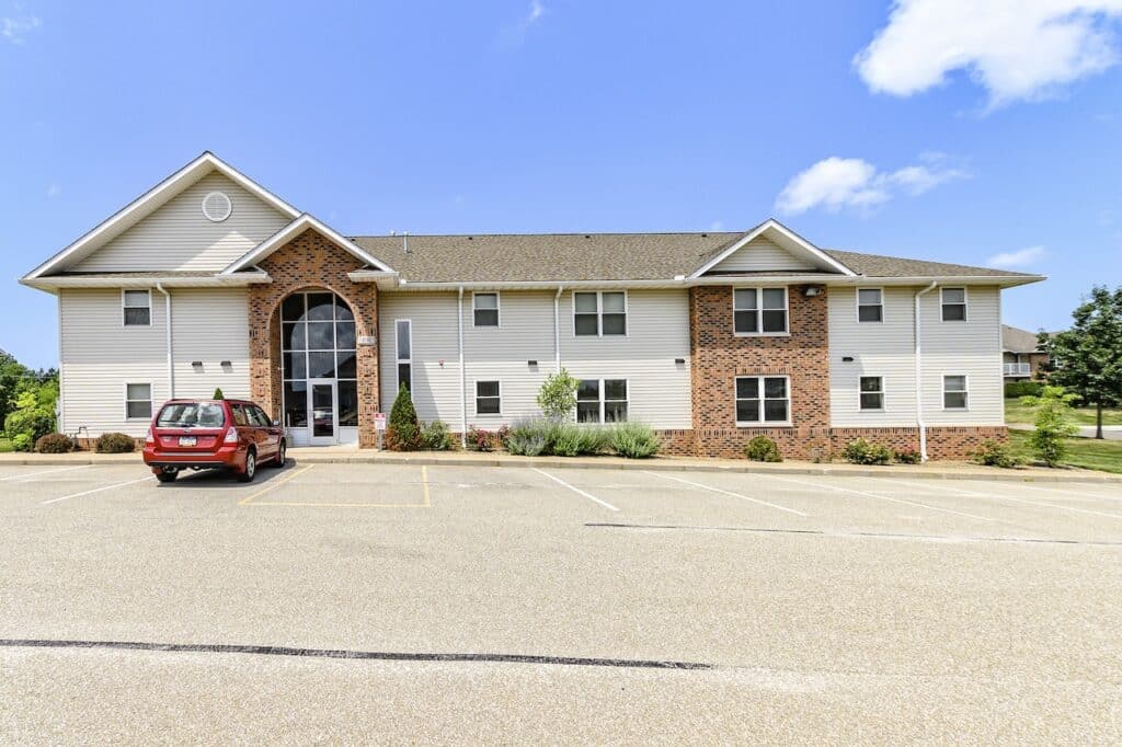 A two-story brick and siding building with a parking lot and a parked red car in front, under a partly cloudy sky.