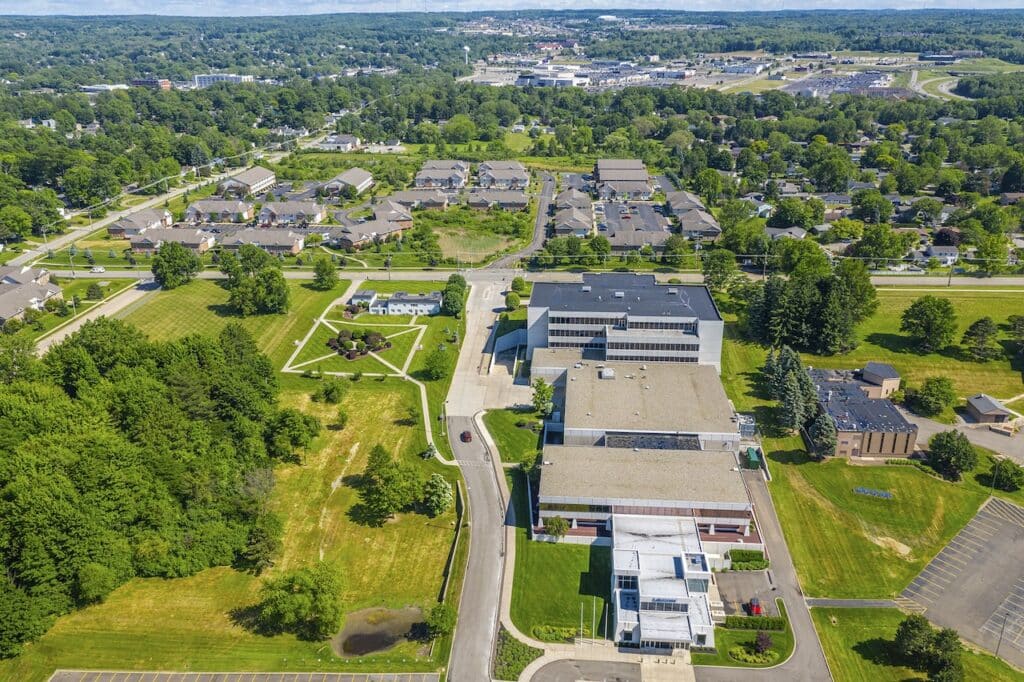 Aerial view of a suburban area with a large modern building, residential houses, roads, and surrounding greenery.