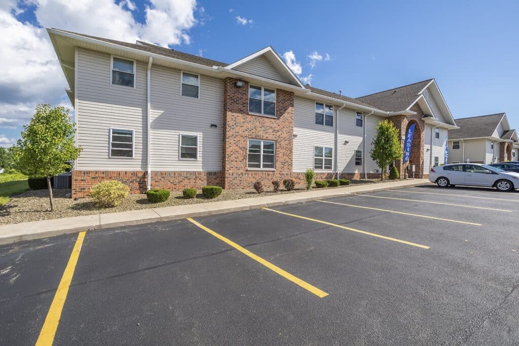 Two-story building with beige siding and brick facade, featuring multiple windows. A parked white car is in the adjacent lot, with clear blue skies overhead.