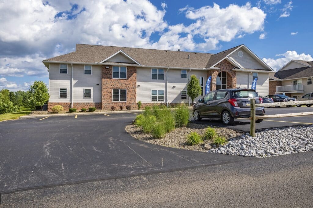 A two-story building with a brick and siding exterior is shown with a parking lot in front. A car is parked near decorative rocks and grass under a partly cloudy sky.