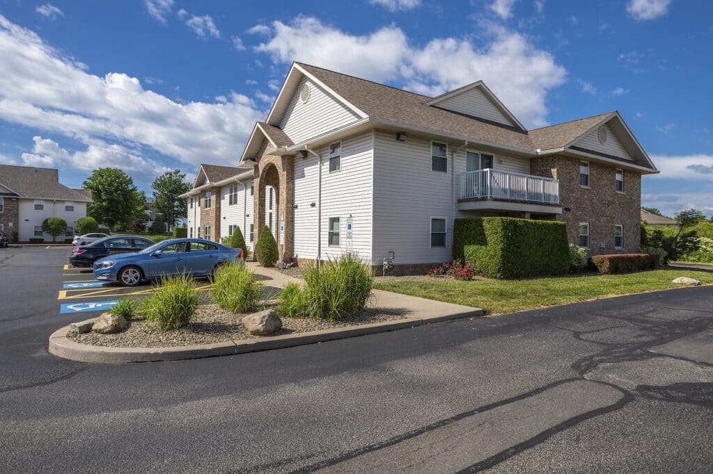 A two-story residential building with tan siding and brick accents, surrounded by a parking lot and landscaped greenery, under a partly cloudy sky.