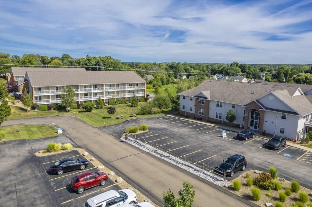 Aerial view of an apartment complex with two beige buildings, a parking lot with parked cars, and surrounding greenery under a blue sky.