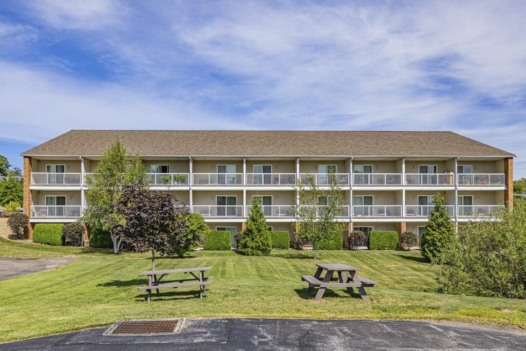 A three-story residential building with balconies, surrounded by green lawns and trees, featuring two picnic tables on the grass in the foreground.