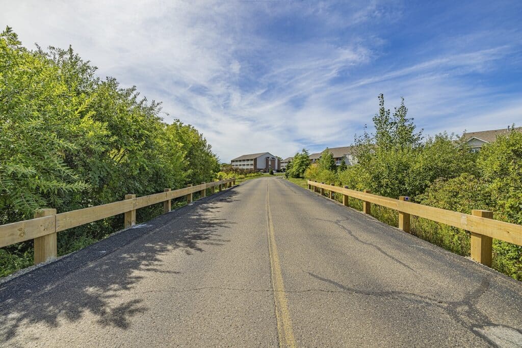 A paved road with a wooden fence on both sides leads to a distant building, surrounded by lush green trees under a clear blue sky.