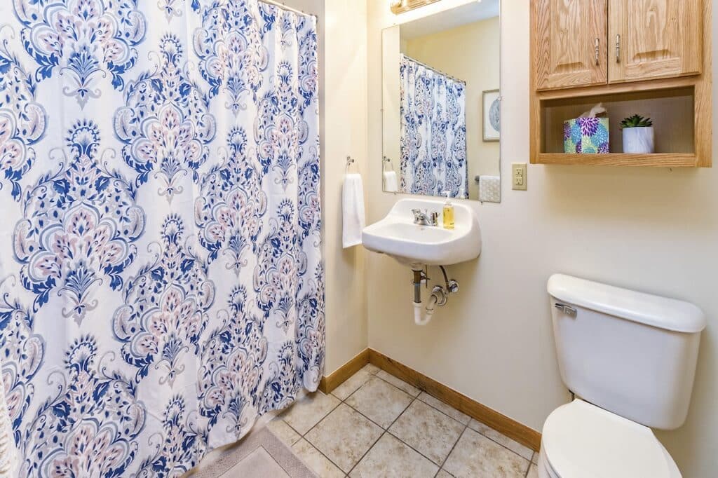 A bathroom with a blue and white floral shower curtain, wall-mounted sink, mirror, wooden cabinet, and toilet. A towel hangs by the sink, and a small plant is on the cabinet.