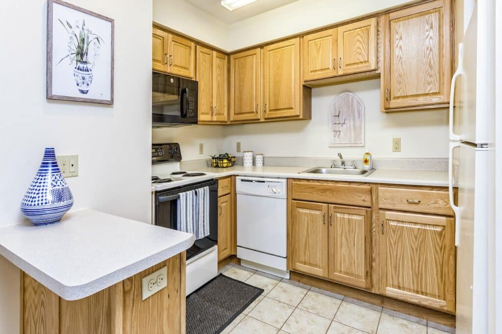 Kitchen with wooden cabinets, white countertops, a white fridge, dishwasher, stove with overhead microwave, and decorative items. Tile flooring and a small counter with an electrical outlet.