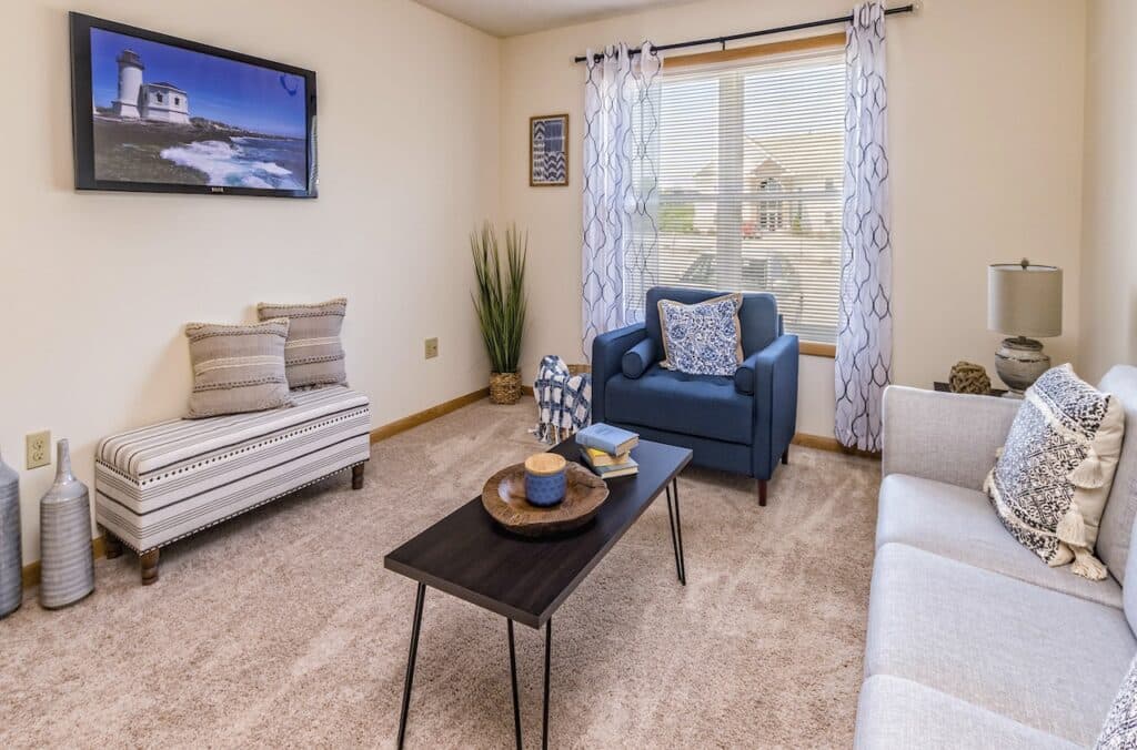 A living room with a blue armchair, white sofa, patterned cushions, a wooden coffee table, and a wall-mounted TV displaying a lighthouse scene. A window with sheer curtains allows natural light in.