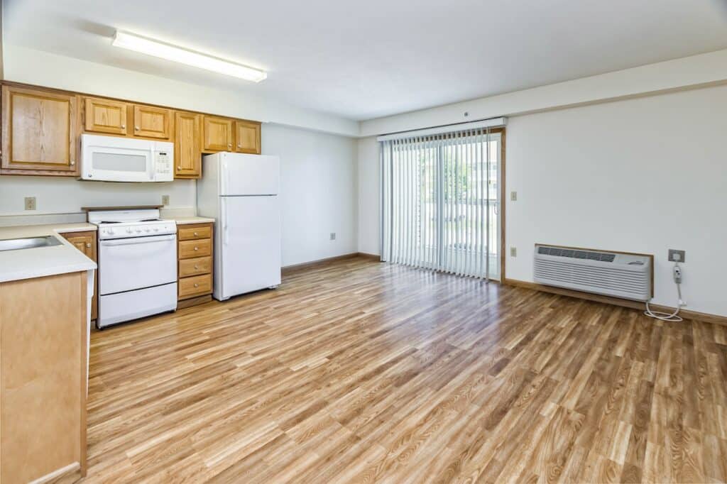 Empty apartment interior featuring a kitchen with wooden cabinets, a white stove, microwave, and refrigerator. The room has wood flooring and sliding glass doors with vertical blinds.
