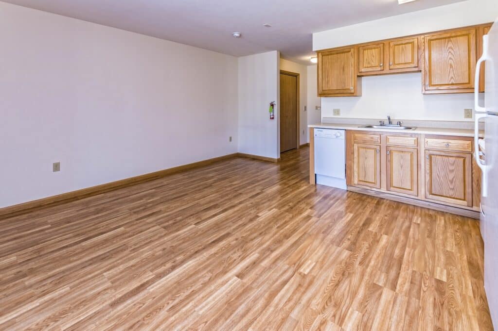 An empty room with wood flooring and a small kitchen area featuring wooden cabinets, a dishwasher, sink, and white fridge on the right. Door to another room is visible in the background.