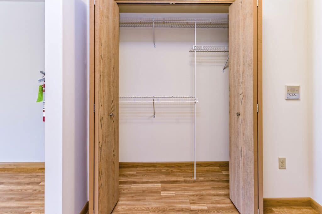 An empty closet with open wooden double doors and white wire shelving, set in a room with light wood flooring.