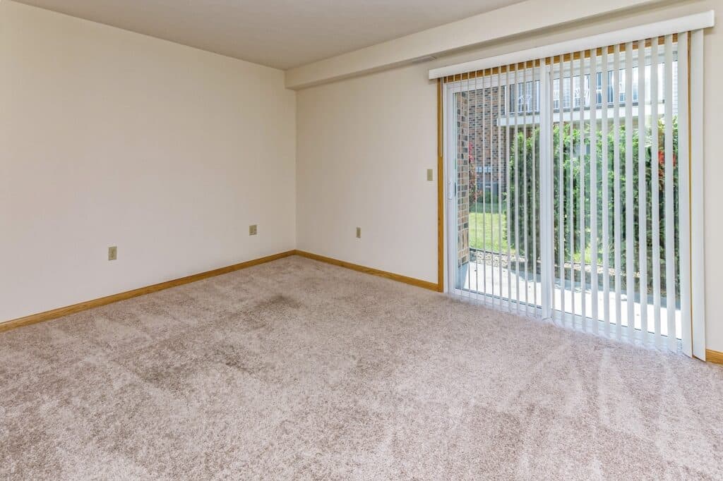 An empty room with beige carpet, white walls, and vertical blinds covering a sliding glass door leading to a garden.