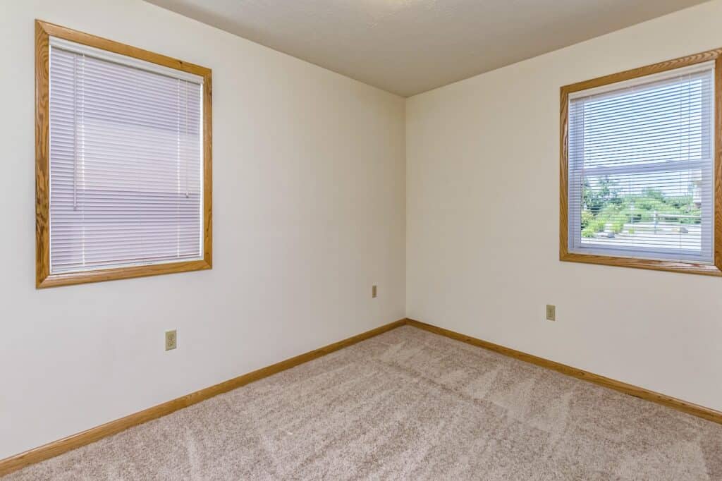Empty room with beige carpet, white walls, and two windows with blinds. The windows have wooden frames and the room is well-lit.