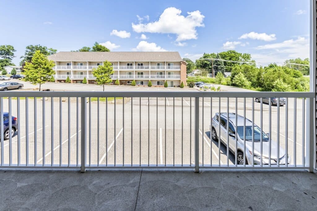 View from a balcony overlooking a parking lot with parked cars. Across the lot is a two-story building with white balconies. The scene is under a clear blue sky.