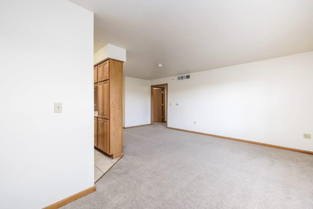 An empty room with beige carpet and white walls, featuring wooden cabinets and a doorway in the background.