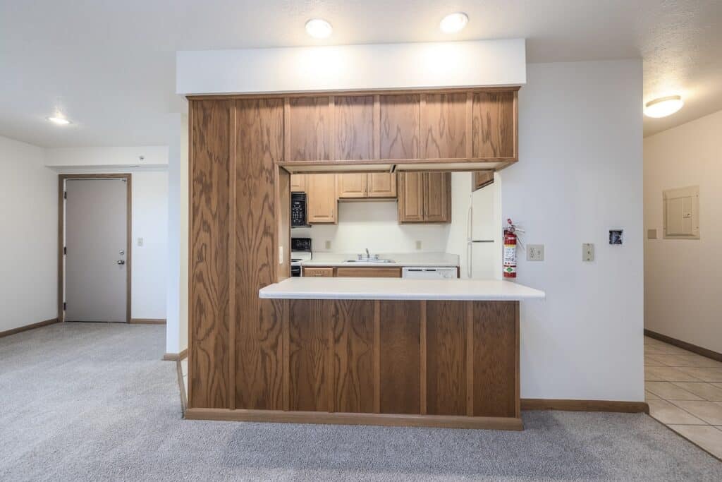 Small kitchen with wood cabinetry, an open counter, and white appliances. There's a carpeted room and a door to the left.