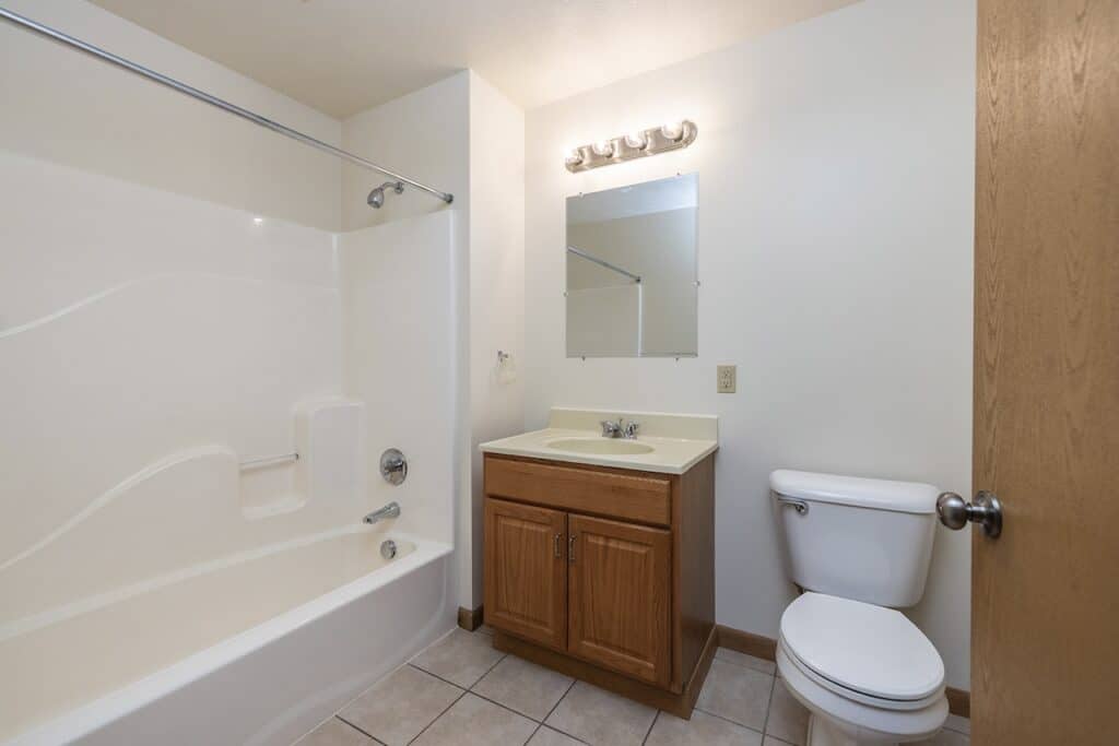 A bathroom with a bathtub-shower combo, a sink with a wooden cabinet, a toilet, and a mirror. Walls are light-colored, and the floor is tiled.