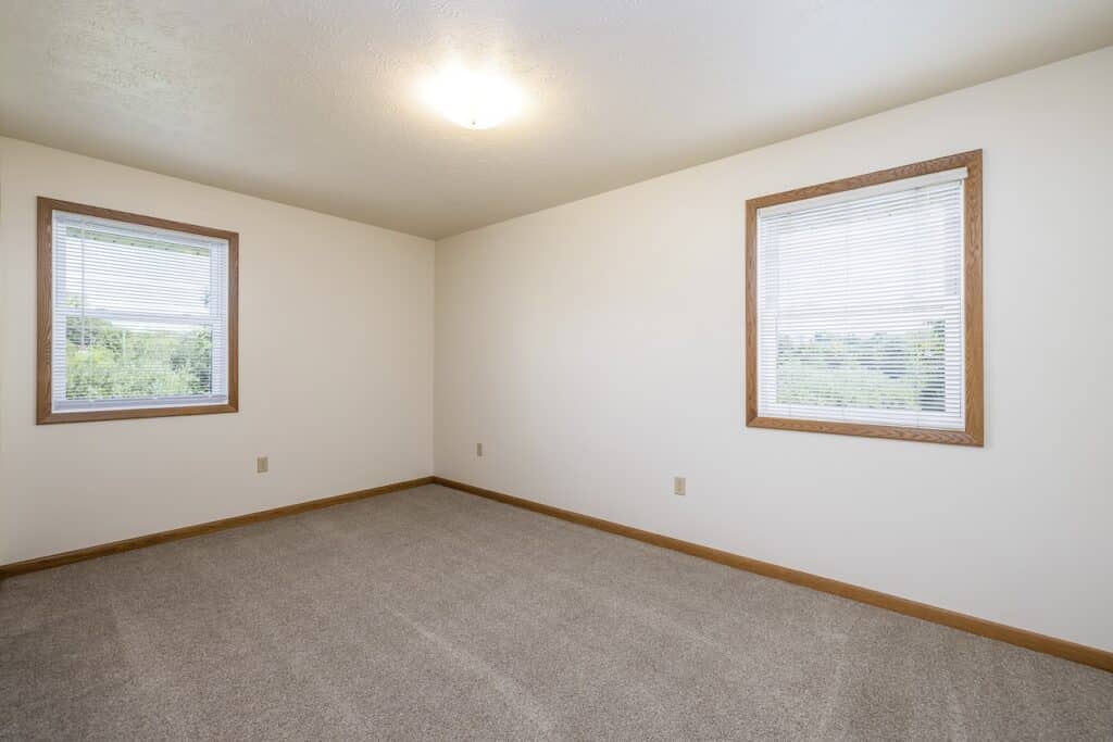 An empty room with light beige walls, beige carpet, and two windows with blinds.