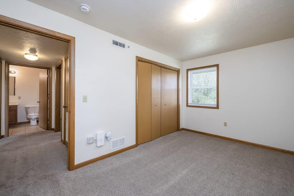 An empty room with beige carpet, wooden closet doors, a window, and a view into a connected bathroom.