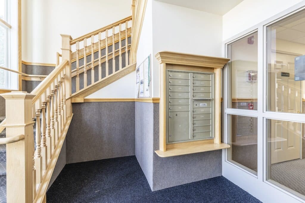 Apartment stairwell with light wood railings, a metal mailbox unit on the wall, and a windowed door on the right.