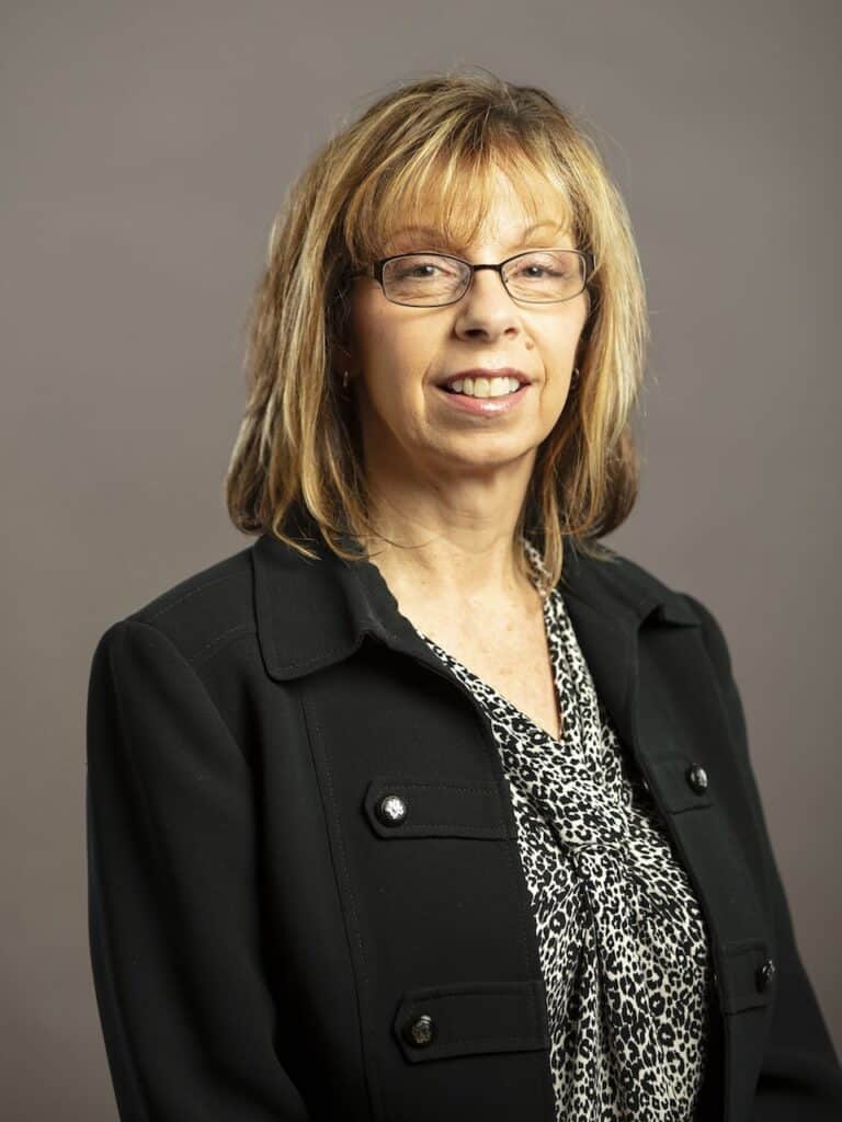 A woman wearing glasses, a black blazer, and a patterned blouse stands against a plain background.