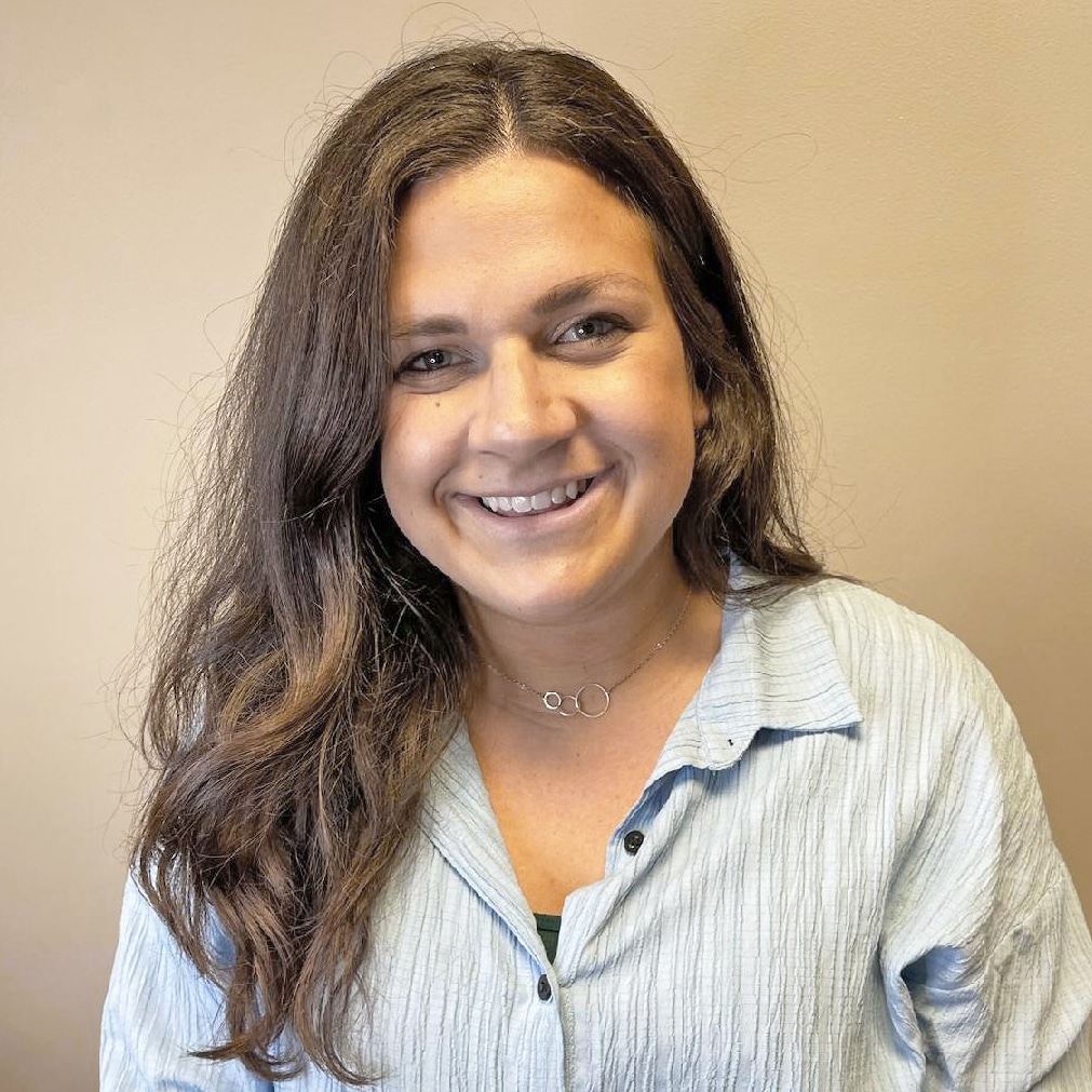 A person with long brown hair smiling, wearing a light blue shirt against a plain beige background.