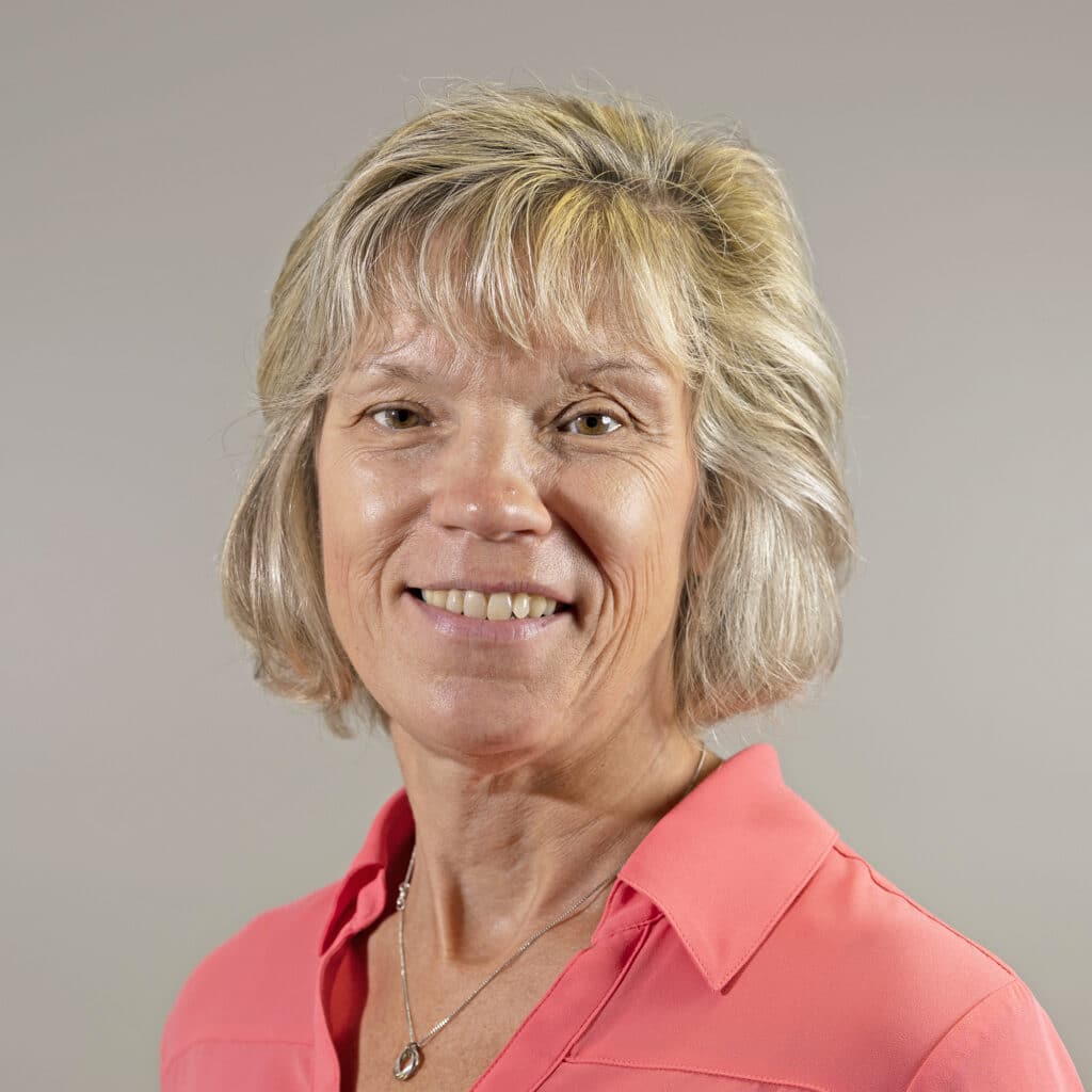 A woman with short blond hair smiles while wearing a coral blouse against a plain background.