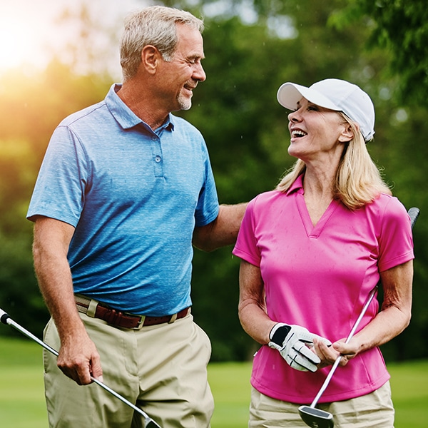 An older man and woman stand outdoors on a golf course, smiling and holding golf clubs. The man wears a blue shirt; the woman wears a pink shirt and white cap.