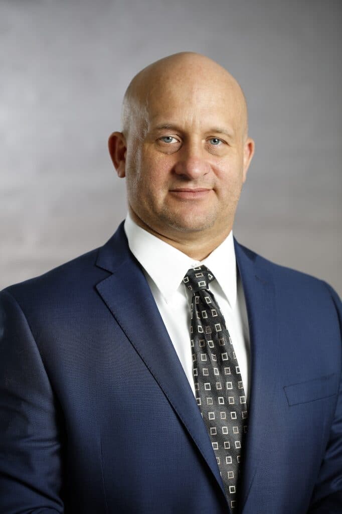 A bald man in a blue suit and patterned tie stands against a gray background, looking directly at the camera.