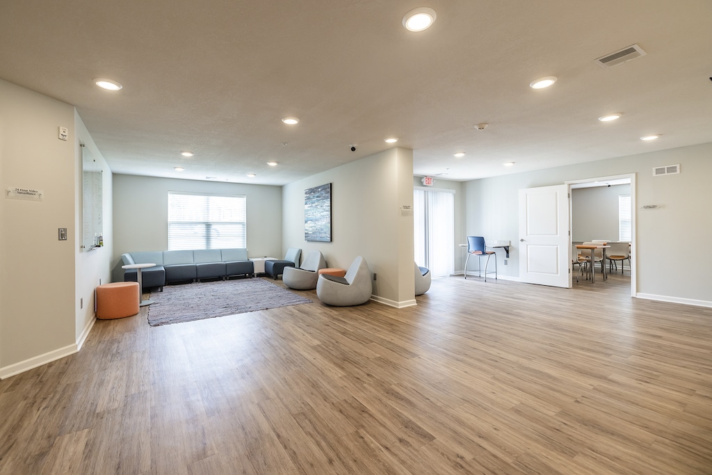 Modern study rooms room with light LVP flooring, gray couches, a rug, and soft lighting. There's a table with chairs in the background near large windows.