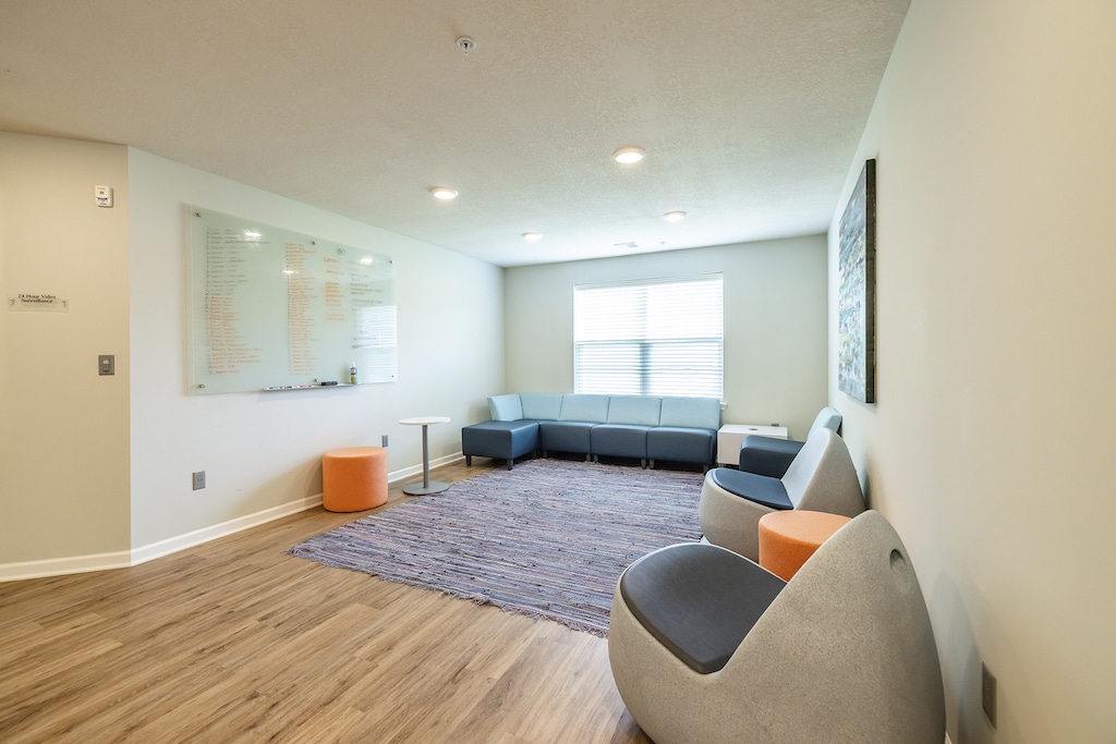 A waiting room with gray chairs, a blue couch, two orange stools, a rug, a window, and a wall-mounted whiteboard.