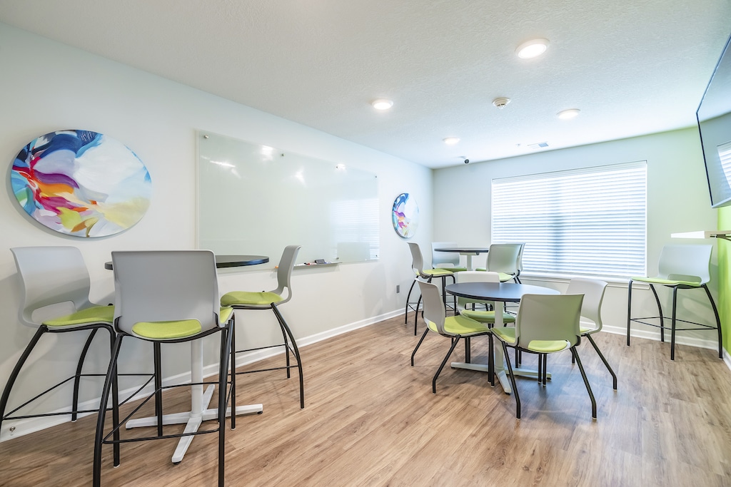 A modern private study with circular tables, high chairs, a large whiteboard, and abstract wall art. Natural light enters through a window with blinds.
