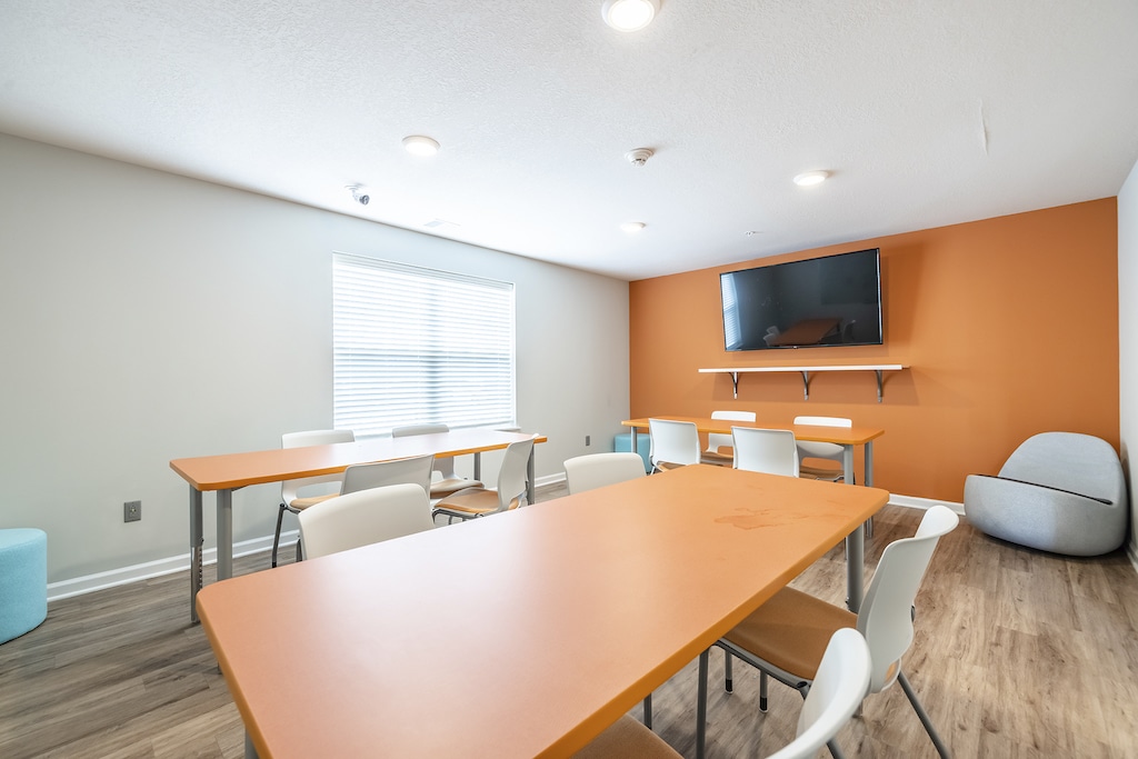 Private study room with orange tables, white chairs, and a TV on an orange accent wall. There's a window with blinds, light LVP flooring, and a modern round gray chair.