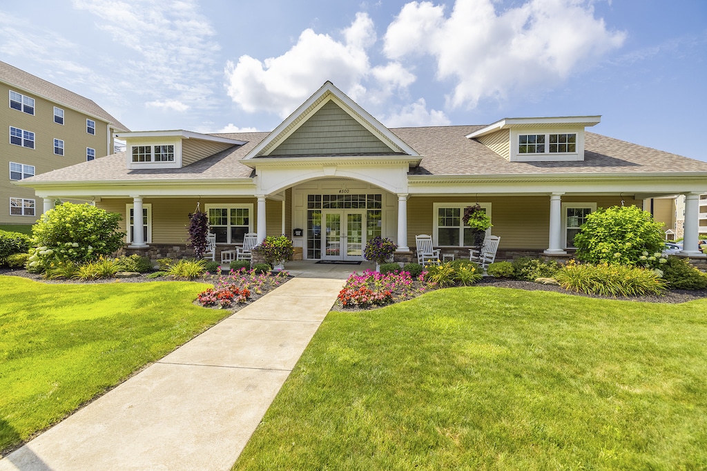 Clubhouse exterior view, single level, with a gabled roof, wide front porch, and manicured garden. Two symmetrical dormers and a pathway lead to the entrance. Bright, sunny day with a few clouds.