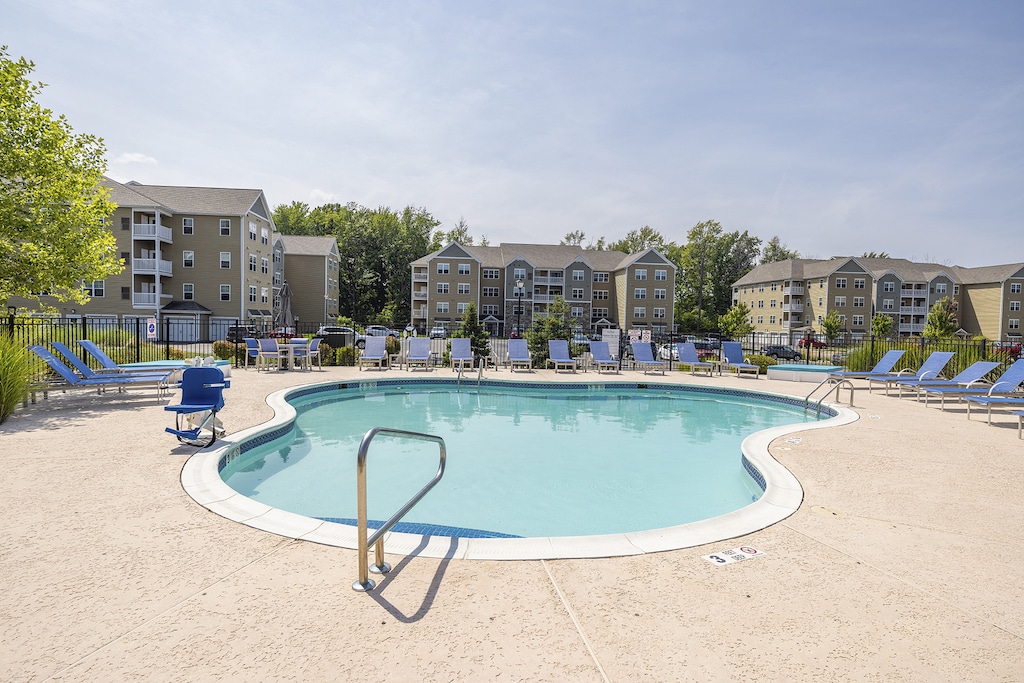 Outdoor swimming pool with a curved shape surrounded by lounge chairs. Apartment buildings and trees are in the background under a clear sky.