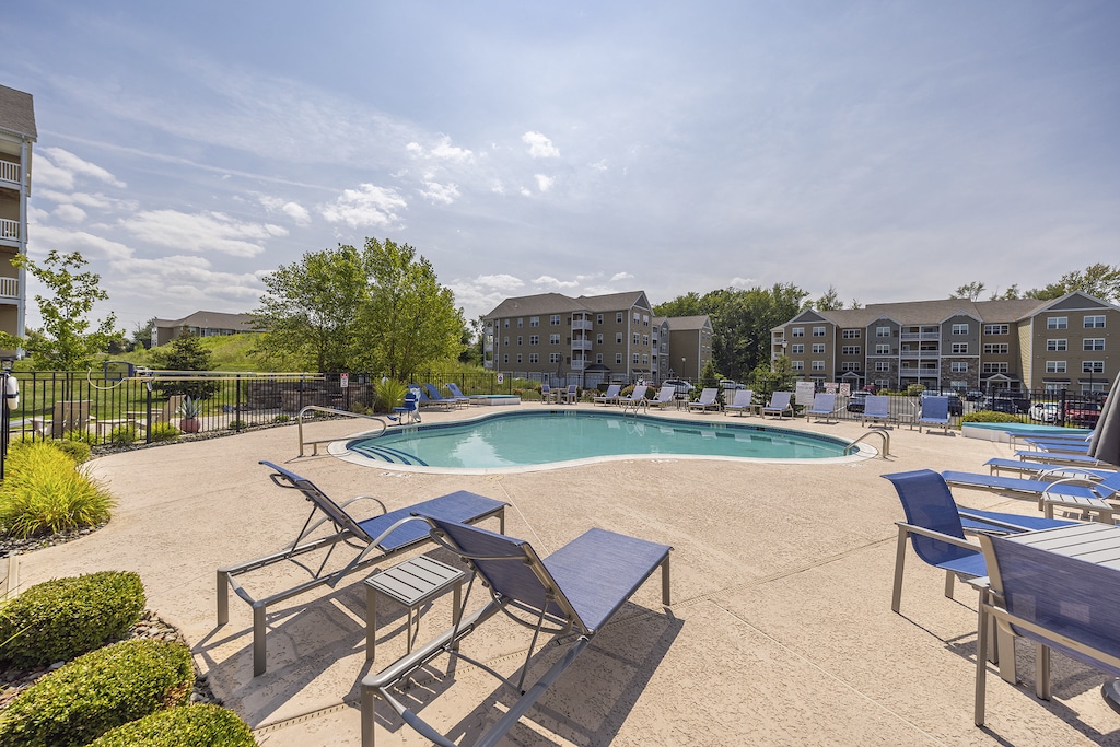 Outdoor apartment complex pool area with lounge chairs, a few trees, and multi-story buildings in the background under a partly cloudy sky.