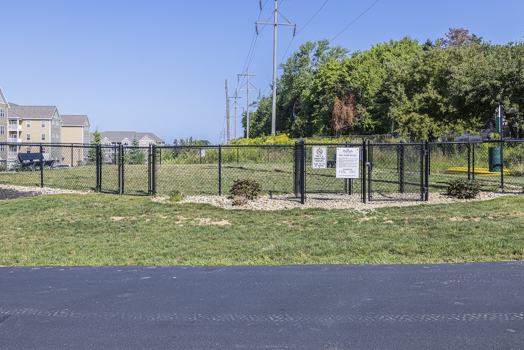 A fenced dog park with signs, surrounded by grass and trees, is located near residential buildings.