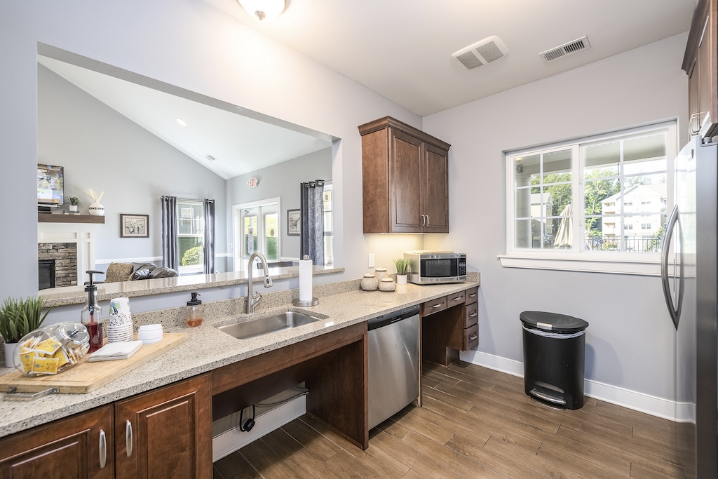 A modern resident kitchen with stainless steel appliances, granite countertops, and wooden cabinets. Large window overlooks the backyard. Adjacent resident lounge area visible through the open wall.