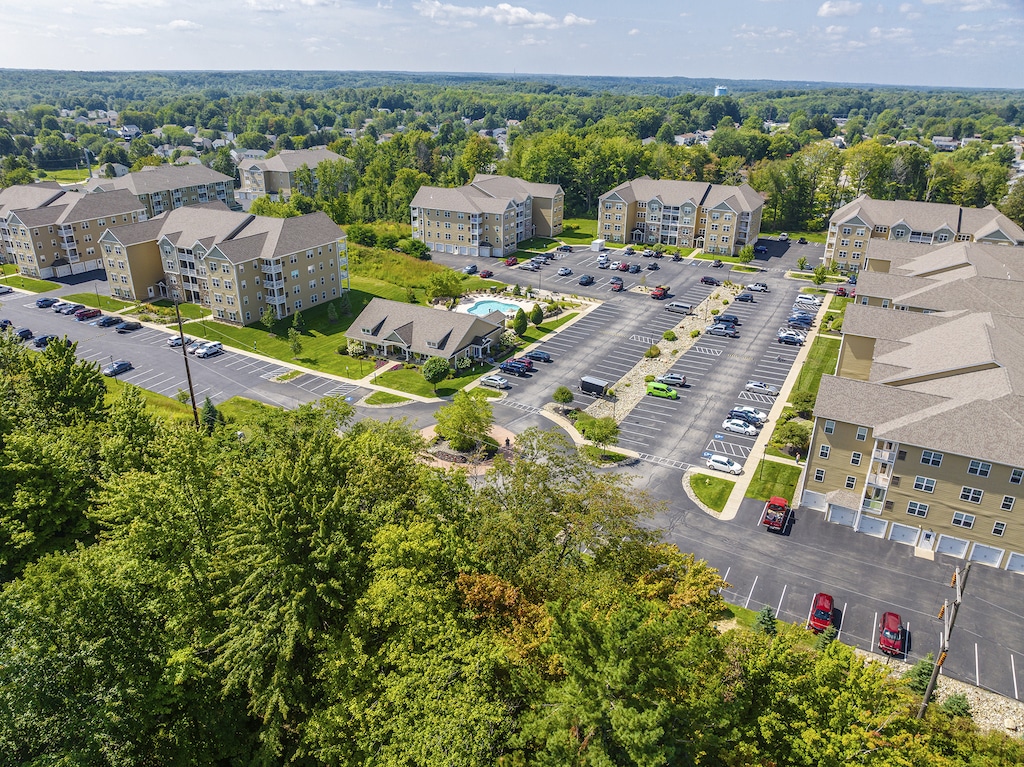 Aerial view of a residential apartment complex with multiple beige buildings, parking lots, greenery, and a swimming pool under a partly cloudy sky.