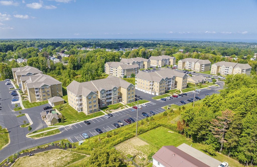 Aerial view of a residential complex with multiple apartment buildings, parking lots, and surrounding greenery under a clear blue sky.