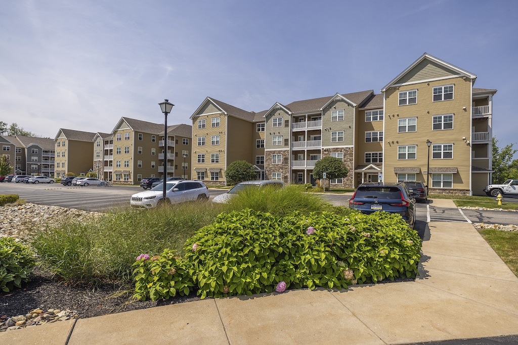 An apartment building with multiple beige buildings, a parking area with cars, and landscaped shrubs in the foreground.