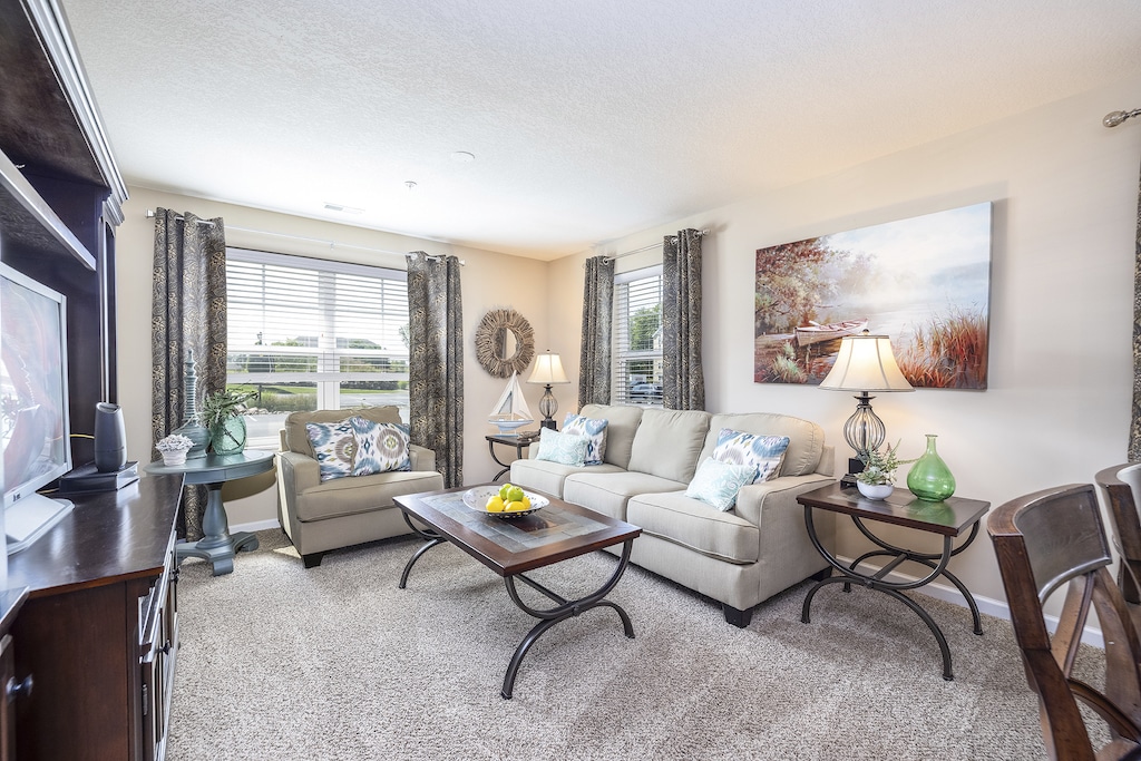 A cozy living room with a beige sofa, armchair, and coffee table. Two windows with curtains, a large wall painting, and a TV are visible. Decorative pillows and lamps add accents.